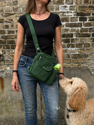 Frau mit schwarzem Oberteil und Jeans trägt eine grüne Nijens Gassi-Tasche mit Flaschenfach, während ein cremefarbener Hund zur Tasche hochschaut.