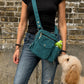 Frau mit schwarzem Oberteil und Jeans trägt eine petrolfarbene Nijens Gassi-Tasche mit Flaschenfach, während ein cremefarbener Hund zur Tasche hochschaut.