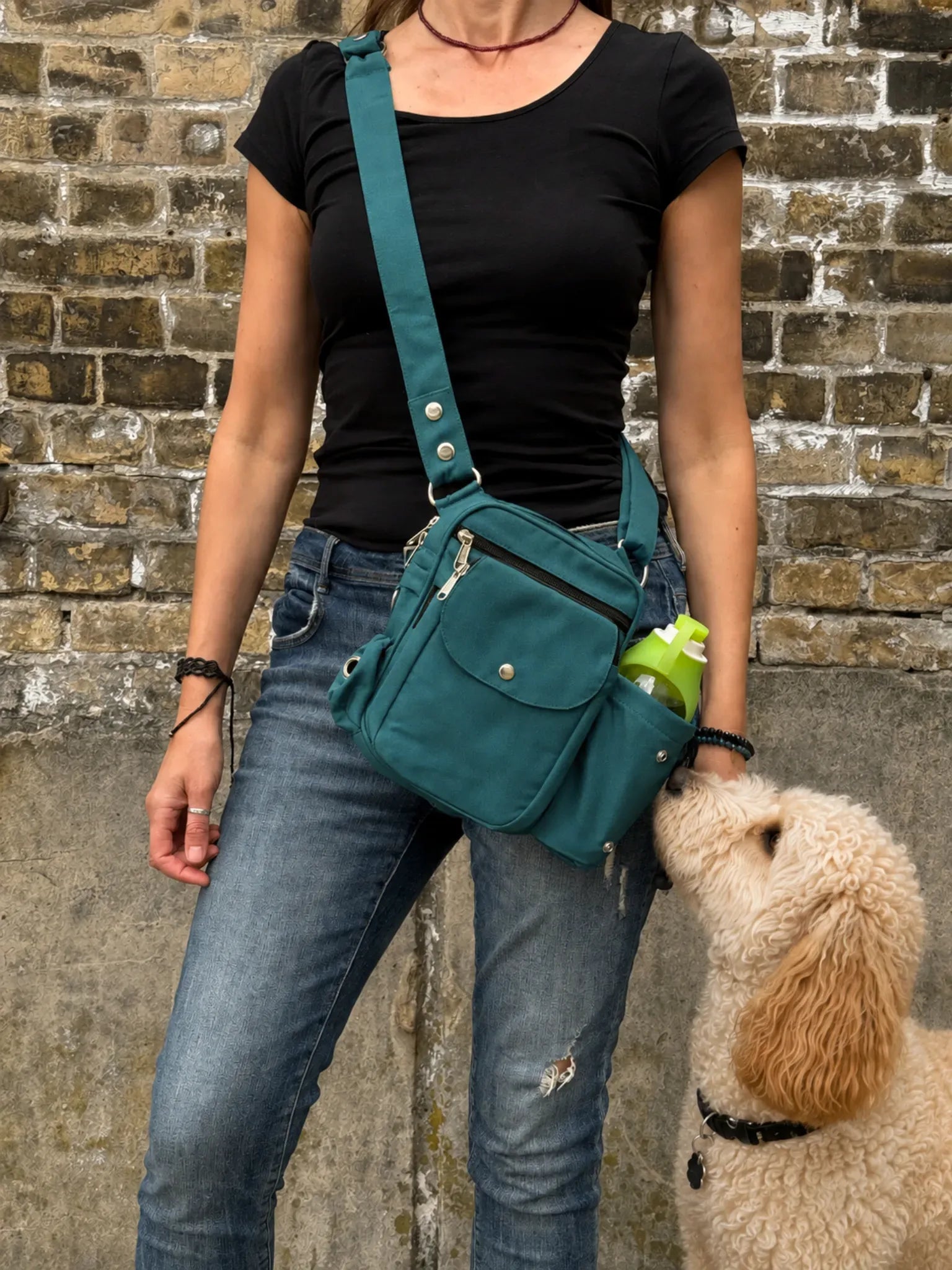 Frau mit schwarzem Oberteil und Jeans trägt eine petrolfarbene Nijens Gassi-Tasche mit Flaschenfach, während ein cremefarbener Hund zur Tasche hochschaut.