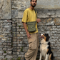Mann mit olivgrüner Crossbody-Tasche im Mandala-Design und sitzendem Hund vor rustikaler Steinmauer.