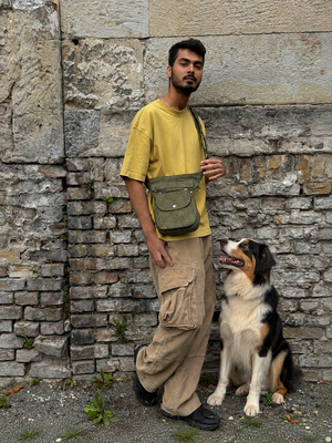 Mann mit olivgrüner Crossbody-Tasche im Mandala-Design und sitzendem Hund vor rustikaler Steinmauer.