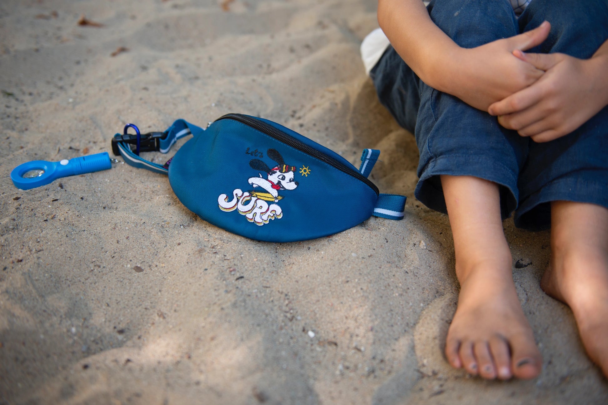 Crossbody Tasche mit Stickmotiv "Let´s SURF" liegt im Sand neben den Füßen des Jungen.