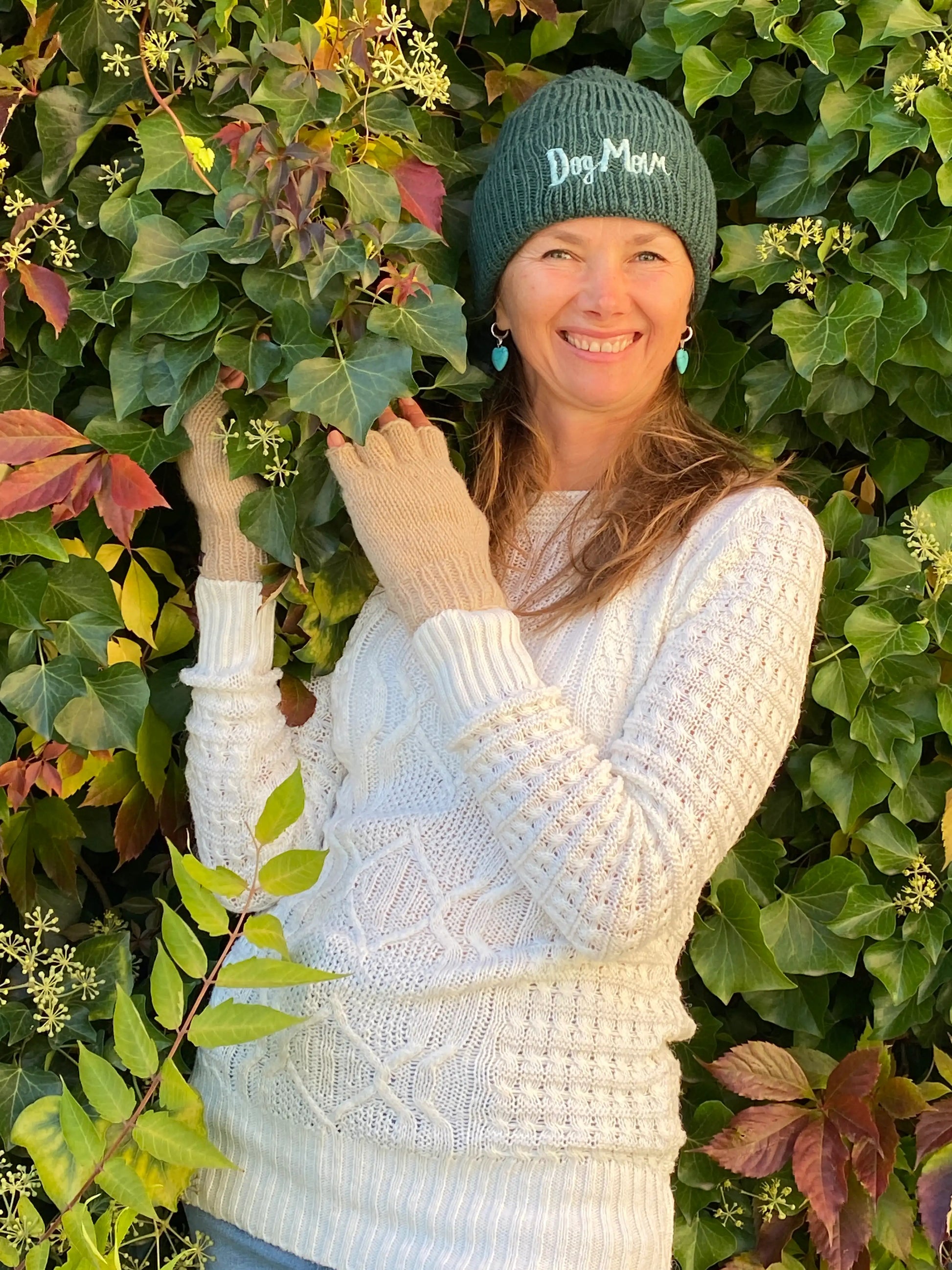 Modell umringt von herbstlichen Blättern und Wollmütze, Strickmütze, Beanie-Mütze aus Schurwolle, Dunkelgrün, mit Umschlag und Fleeceeinsatz. Der Umschlag ist vorne mit dem Schriftzug „Dog Mom“ bestickt.