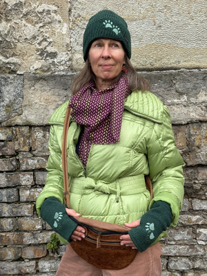 Model präsentiert eine dunkelgrüne Beanie-Mütze und die dazu passenden Handstulpen mit Daumenschlitz aus Schurwolle, bestickt mit zwei Pfoten-Logo.