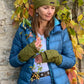 Model präsentiert  in herbstlicher Kulisse die kurze Beanie-Mütze, Strickmütze aus Schurwolle, Grün, bestickt mit Pfote-Herz-Logo und Handstulpen aus Schurwolle, Grün, mit Fingerkappe.