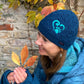 Seitenansicht: Model präsentiert die kurze Beanie-Mütze, Strickmütze aus Schurwolle, Petrol, bestickt mit Pfote-Herz-Logo und Handstulpen aus Schurwolle, Petrol, mit Fingerkappe.