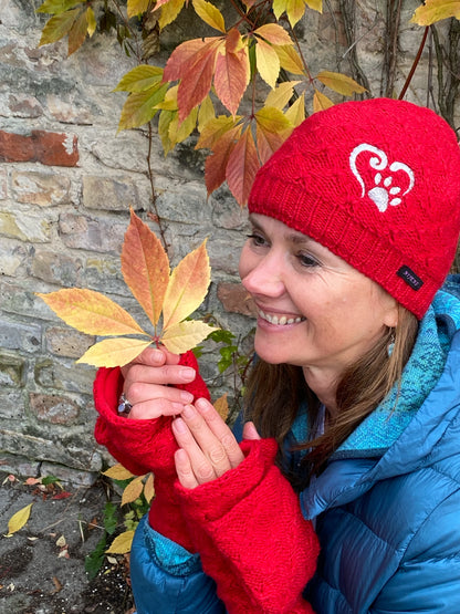 Model präsentiert die kurze Beanie-Mütze, Strickmütze aus Schurwolle, Rot, bestickt mit Pfote-Herz-Logo an der Seite und Handstulpen aus Schurwolle Rot mit Fingerkappe.