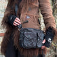 Model präsentiert Gassi-Tasche Crossbody-Bag, Bauchtasche Berlin aus Baumwoll-Canvas schwarz mit floralen Ornamenten. Tragevariante: Umhängetasche. An den Händen trägt sie Armstulpen aus Schurwolle, schwarz bestickt mit zwei Pfoten-Logo.