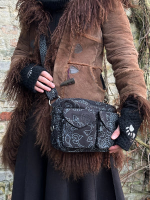 Model präsentiert Gassi-Tasche Crossbody-Bag, Bauchtasche Berlin aus Baumwoll-Canvas schwarz mit floralen Ornamenten. Tragevariante: Umhängetasche. An den Händen trägt sie Armstulpen aus Schurwolle, schwarz bestickt mit zwei Pfoten-Logo.