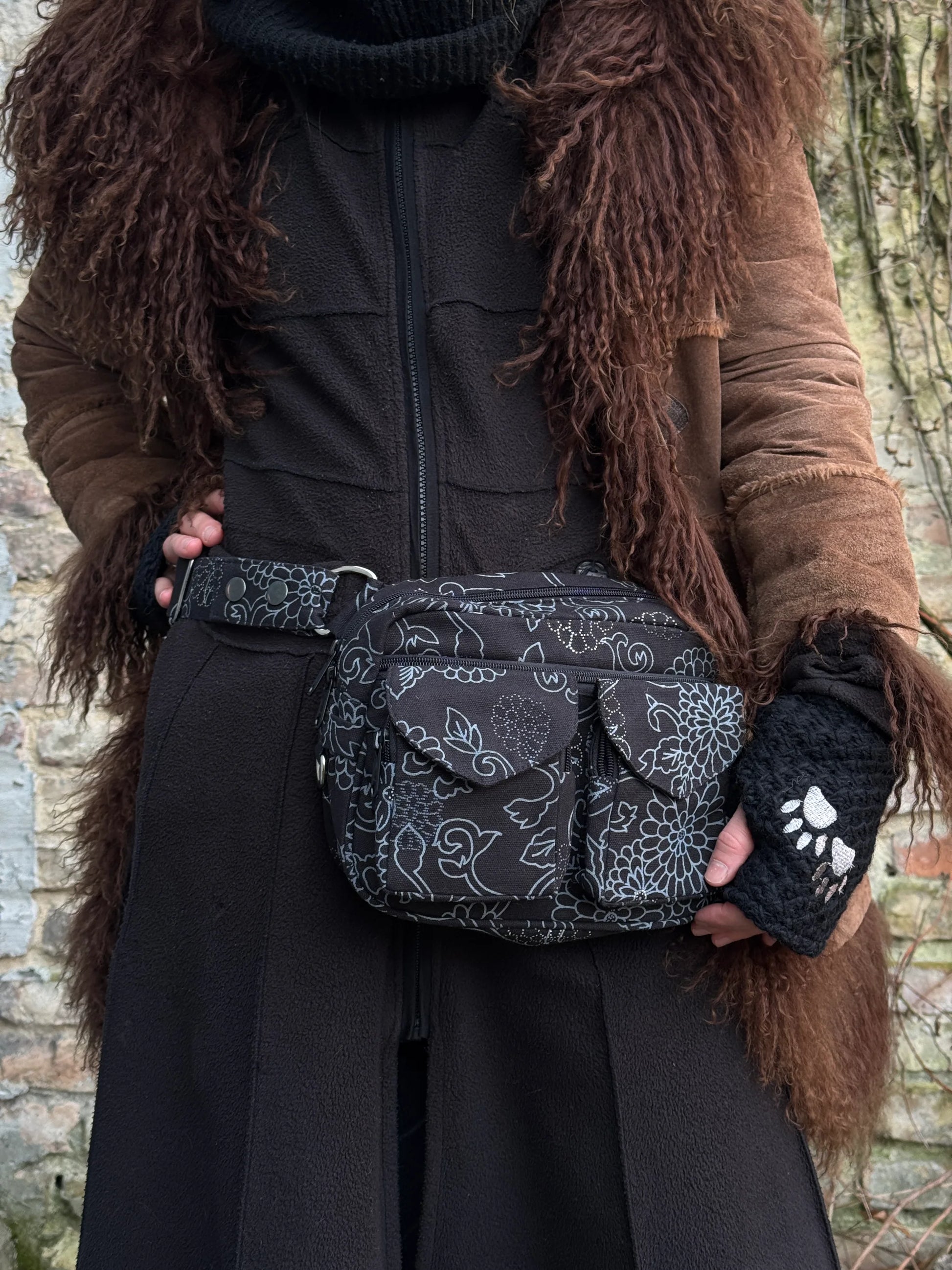 Model präsentiert Gassi-Tasche Crossbody-Bag, Bauchtasche Berlin aus Baumwoll-Canvas schwarz mit floralen Ornamenten. Tragevariante: Hüfttasche. An den Händen trägt sie Armstulpen aus Schurwolle, schwarz bestickt mit zwei Pfoten-Logo.