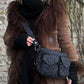 Model präsentiert Gassi-Tasche Crossbody-Bag, Bauchtasche Berlin aus Baumwoll-Canvas schwarz mit kleinem Pfoten-Print. Tragevariante: Umhängetasche. An den Händen trägt sie dezent gestreifte Armstulpen aus Schurwolle in Schwarz und Anthrazit.