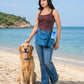 Frau mit blauer Cord-Gassitasche und integriertem Flaschenhalter neben einem Golden Retriever am Strand mit blauem Meer im Hintergrund.