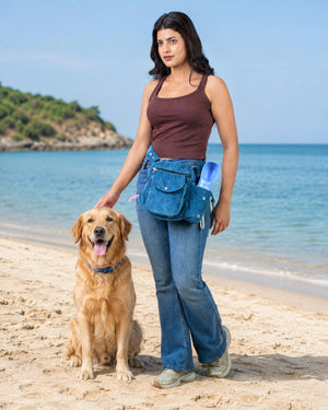 Frau mit blauer Cord-Gassitasche und integriertem Flaschenhalter neben einem Golden Retriever am Strand mit blauem Meer im Hintergrund.