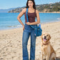 Frau mit blauer Cord-Tasche und integriertem Flaschenhalter neben einem Golden Retriever am Strand mit blauem Meer im Hintergrund.