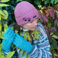 Model vor herbstlicher Kulisse präsentiert handgestrickte Handschuhe und kurze Wollmüzue aus Schurwolle in Türkis/ Rosa mit Fleeceinlage und Zopfstrickmuster auf dem Handrücken.