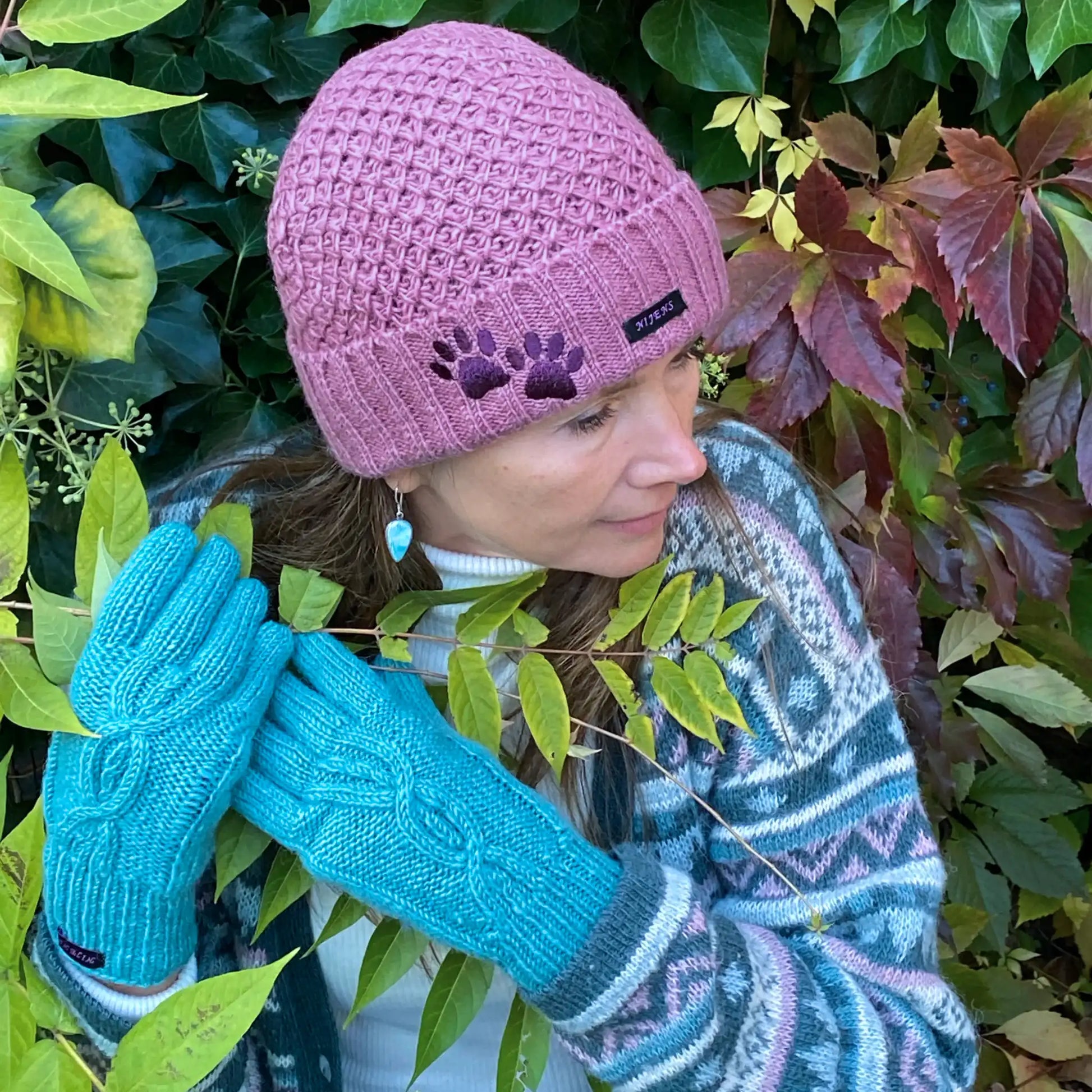 Model vor herbstlicher Kulisse präsentiert handgestrickte Handschuhe und kurze Wollmüzue aus Schurwolle in Türkis/ Rosa mit Fleeceinlage und Zopfstrickmuster auf dem Handrücken.