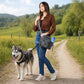 Frau mit grauer Nijens Gassitasche und weißen Sportschuhen steht mit Husky auf einem Feldweg in grüner Landschaft.