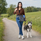 Frau mit grauer Gassitasche von Nijens und Sportschuhen spaziert mit Husky auf einem Weg durch ein Feld.