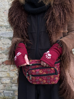 Nahaufnahme: Model mit Crossbody-Gassi-Tasche aus Baumwoll-Canvas in Rot mit bunten, großen Pfoten-Prints. Tragevariante Bauchtasche. Das Model trägt rote Armstulpen aus Schurwolle, bestickt mit 2-Pfoten-Logo.