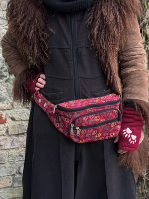 Model präsentiert Crossbody-Gassi-Tasche aus Baumwoll-Canvas in Rot mit bunten, großen Pfoten-Prints. Tragevariante Bauchtasche. Das Model trägt rote Armstulpen aus Schurwolle, bestickt mit 2-Pfoten-Logo.