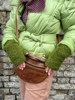 Model präsentiert handgestrickte Handstulpen mit Daumen aus Schurwolle in Olivgrün mit Fleeceeinsatz und Strickmuster. Um die Schulter trägt sie die Crossbody-Bag Bangkok aus Leder mit floralen Ornamentenprägung.