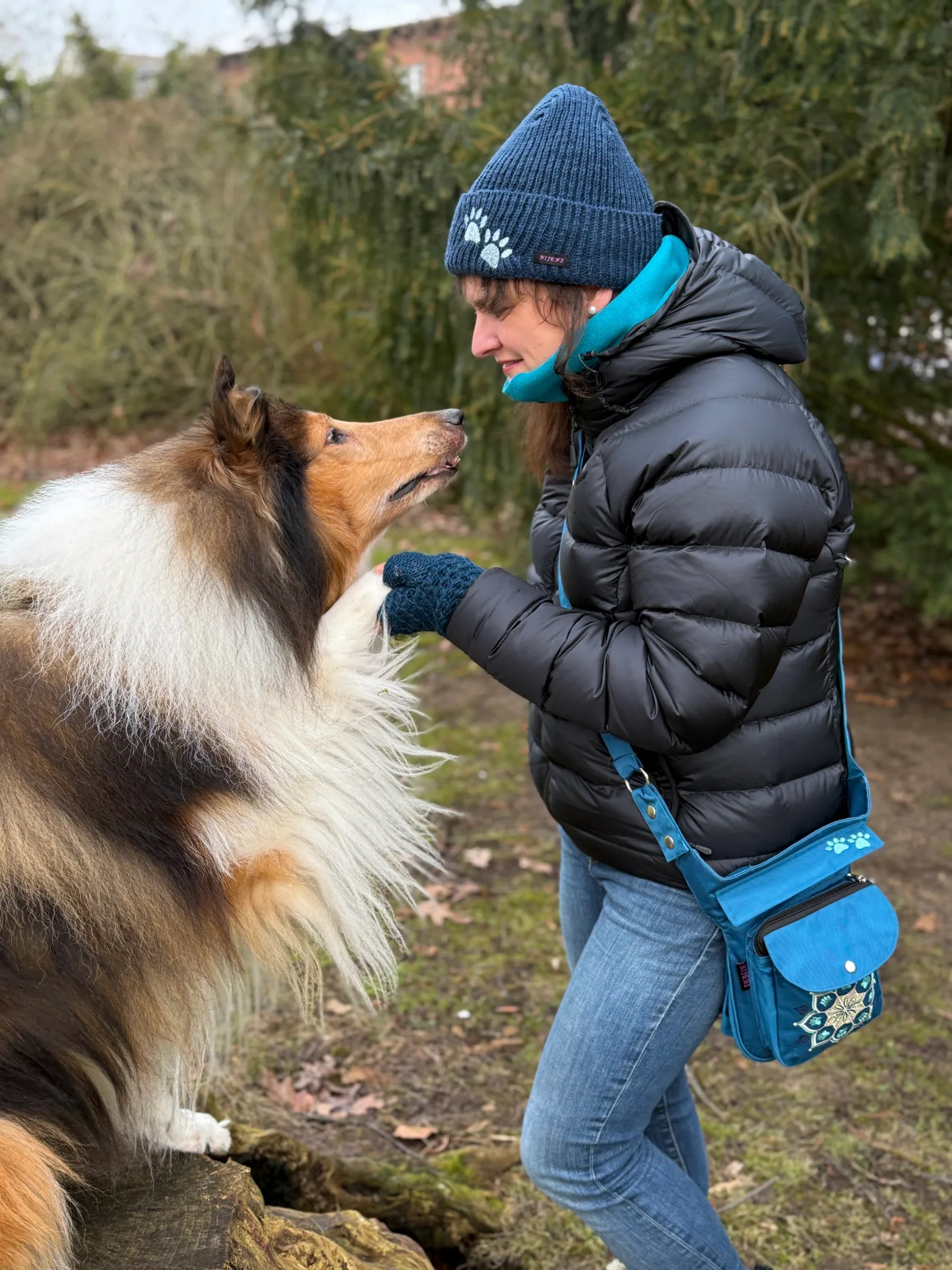 Frau im Park mit Collie-Hund trägt eine türkise Nijens Gassitasche mit Mandala-Stickerei, blaue Strickmütze und Armstulpen.