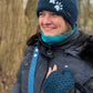Portrait einer Frau im Park mit dunkelblauer Strickmütze mit Pfotenmotiv, Schal und blauen Armstulpen; Nijens Tragegurt sichtbar.