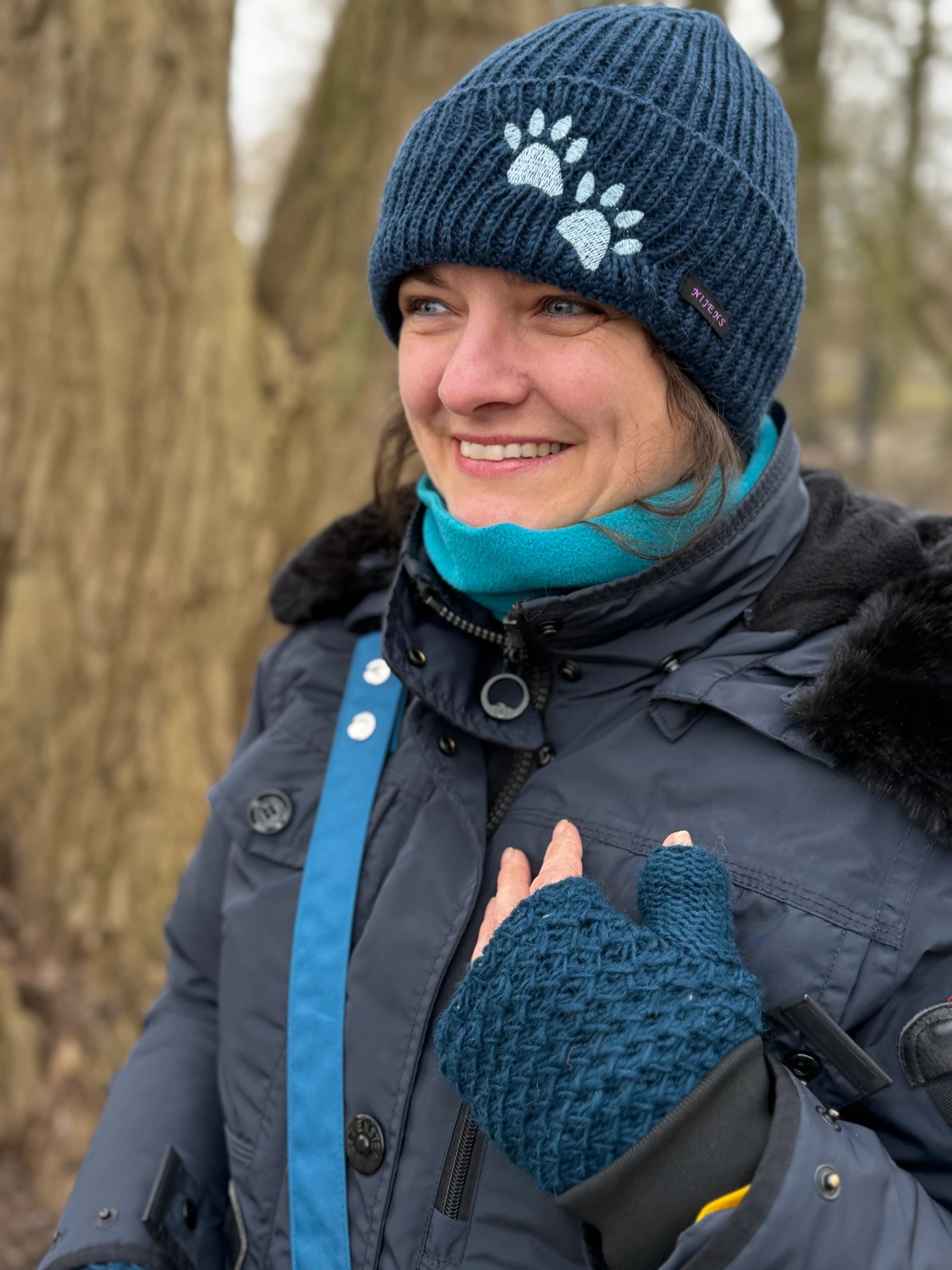 Portrait einer Frau im Park mit dunkelblauer Strickmütze mit Pfotenmotiv, Schal und blauen Armstulpen; Nijens Tragegurt sichtbar.