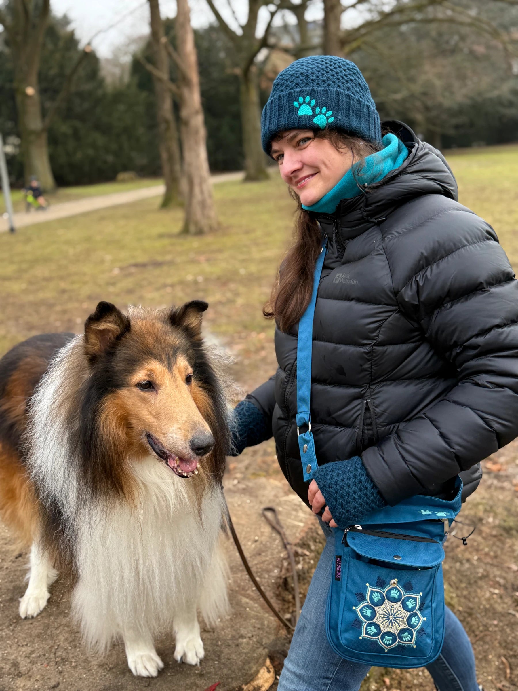 Frau im Park mit Collie-Hund trägt eine türkise Gassitasche mit Mandala-Stickerei, blaue Strickmütze und Armstulpen.