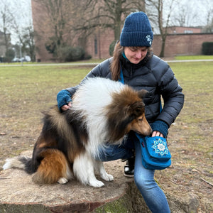 Frau im Park mit blauer Nijens-Umhängetasche mit Mandala-Motiv gibt einem Collie ein Leckerli; sie trägt Mütze und Handschuhe, der Hund sitzt auf einem Baumstumpf.