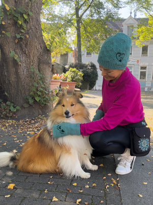 Frau mit türkisfarbener Wollmütze und passenden Pulswärmern von Nijens beim Spaziergang mit ihrem Hund im Herbst, trägt schwarze Gassitasche mit Mandala-Stickerei.