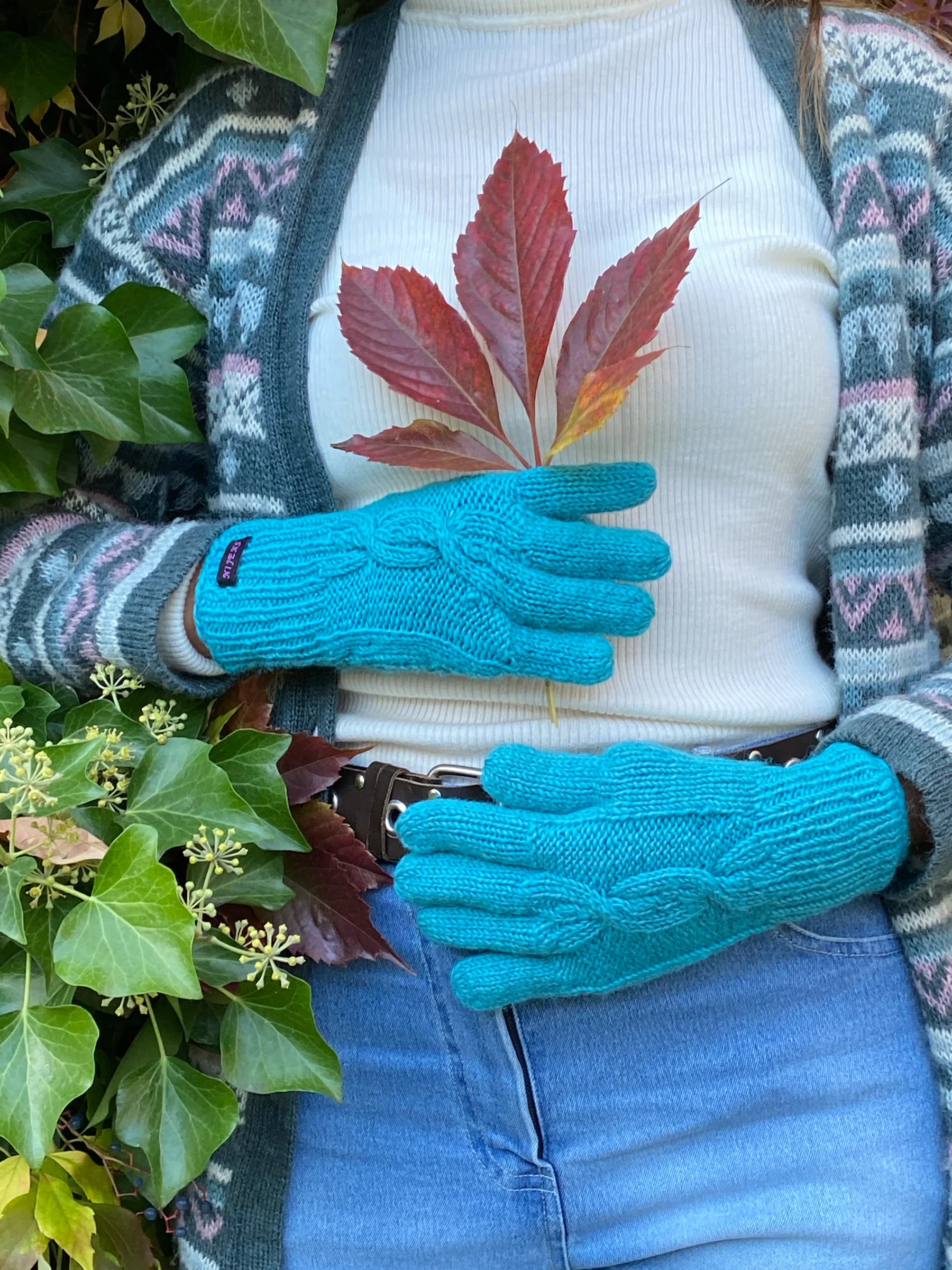 Handmodel vor herbstlicher Kulisse präsentiert handgestrickte Handschuhe aus Schurwolle in Türkis mit Fleeceinlage und Zopfstrickmuster auf dem Handrücken.