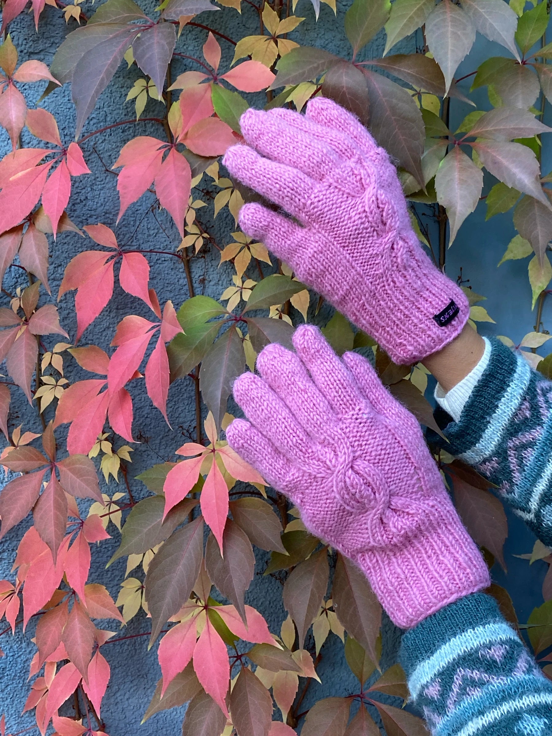 Handmodell präsentiert handgestrickte Handschuhe aus Schurwolle in Rosa mit Fleeceinlage und Strickmuster vor herbstlichen Blättern.