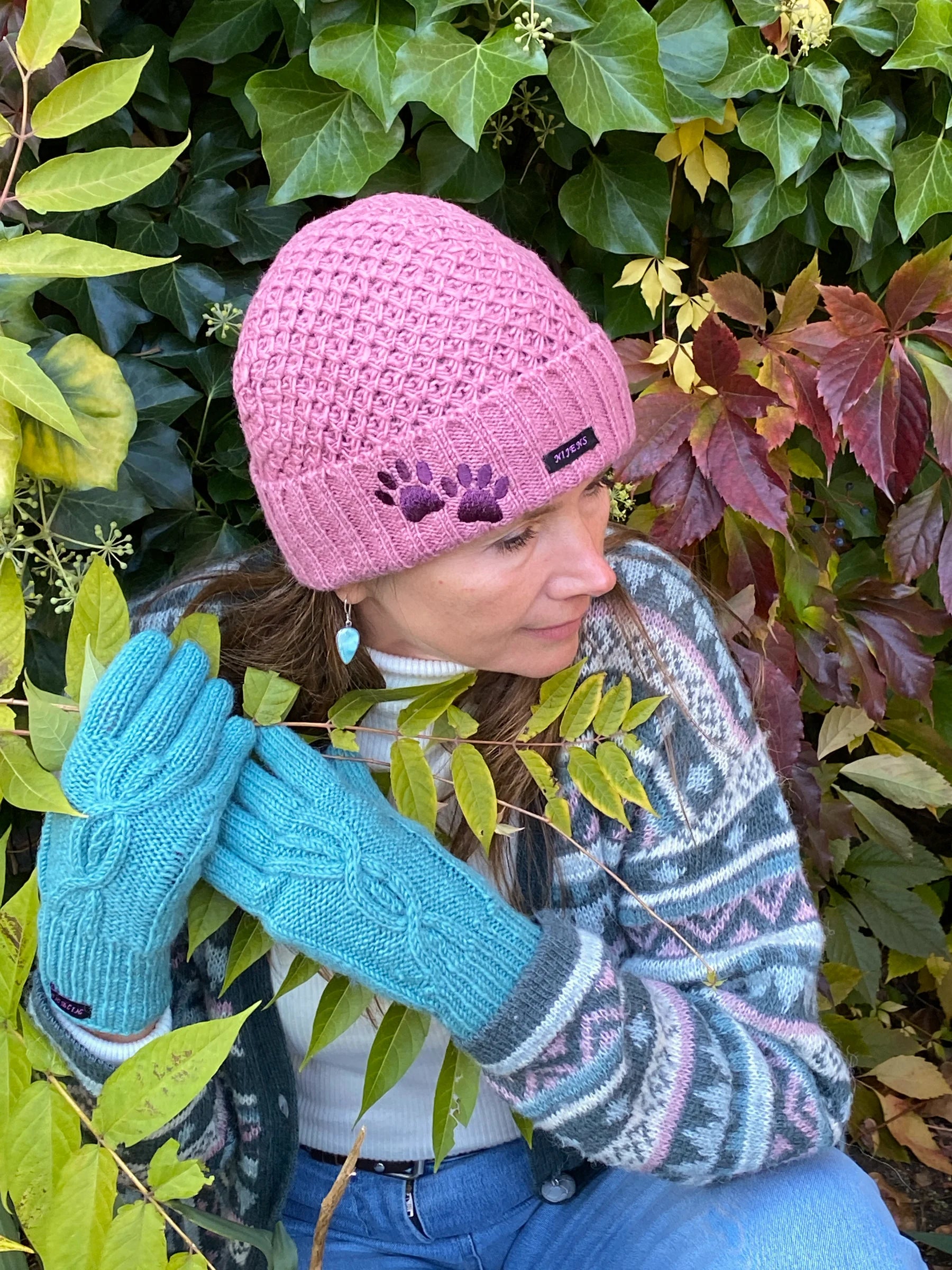 Model vor herbstlicher Kulisse präsentiert handgestrickte Handschuhe und kurze Wollmüzue aus Schurwolle in Türkis/ Rosa mit Fleeceinlage und Zopfstrickmuster auf dem Handrücken.