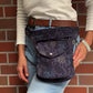 Eine Frau präsentiert eine Gassi-Tasche, Hüfttasche aus robustem Baumwollstoff in Violett mit floralen Ornamenten. 