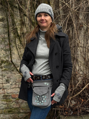 Ein Model trägt auf ihrem Kopf Nijens kurze Beanie-Mütze MINILOONNA 21 aus Schurwolle, Grau, bestickt mit Pfote-Herz-Logo. An den Händen trägt sie Handstulpen HADAR 21 aus Schurwolle in grau mit zwei Pfotenstickerei. Um die Hüfte präsentiert das Model die Bauchtasche Barcelona Mandala 21 aus Baumwoll-Canvas in grau.
