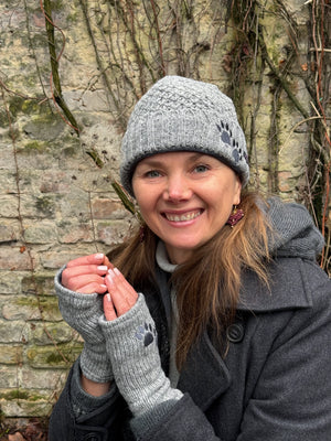 Nahaufnahme: Model trägt kurze Beanie-Mütze MINILOONNA 21 aus Schurwolle, Grau, bestickt mit Pfote-Herz-Logo. An den Händen trägt sie Handstulpen HADAR 21 aus Schurwolle in grau mit zwei Pfotenstickerei.