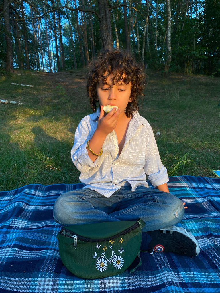 Ein Junge sitzt mit gekreuzten Beinen im Wald auf einem Waldboden und isst Apfel, vor ihm liegt eine Crossbody-Tasche mit Stickmotiv "Schmetterling Fahrrad". 