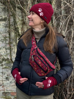 Model präsentiert Crossbody-Gassi-Tasche aus Baumwoll-Canvas in Rot mit bunten, großen Pfoten-Prints. Tragevariante Umhängetasche. Das Model trägt eine rote Beanie-Mütze und rote Armstulpen aus Schurwolle, bestickt mit 2-Pfoten-Logo.