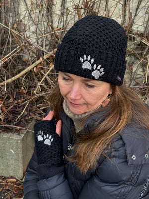 Model präsentiert eine Beanie-Mütze und die dazu passenden Handstulpen mit Daumenschlitz aus schwarzer Schurwolle. Wollmütze und Handstulpen sind bestickt mit einem zwei Pfoten-Logo.