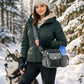 Sportliche Frau in dunkelgrüner Winterjacke, beigem Hoodie, Angora-Mütze und schwarzen Armstulpen steht mit einem Husky in einem verschneiten Wald und trägt die Nijens Gassi-Tasche Konstanz 4711 in Grau aus Canvas und Leder mit Pfoten-Herz-Prägung, Metalldruckknopf und Flaschenhalter auf der rechten Seite.