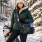 Sportliche Frau in dunkelgrüner Winterjacke, beigem Hoodie, Angora-Mütze und schwarzen Armstulpen steht mit einem Husky in einem verschneiten Wald und trägt die Nijens Gassi-Tasche Konstanz 4716 in Schwarz aus Canvas und Leder mit Pfoten-Herz-Prägung, Metalldruckknopf und Flaschenhalter auf der rechten Seite.