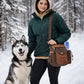 Sportliche Frau in dunkelgrüner Winterjacke und beigem Hoodie steht mit einem Husky in einem verschneiten Wald und trägt die Nijens Gassi-Tasche Konstanz 4718 aus braunem Canvas und Leder mit Pfoten-Herz-Prägung und Flaschenhalter.