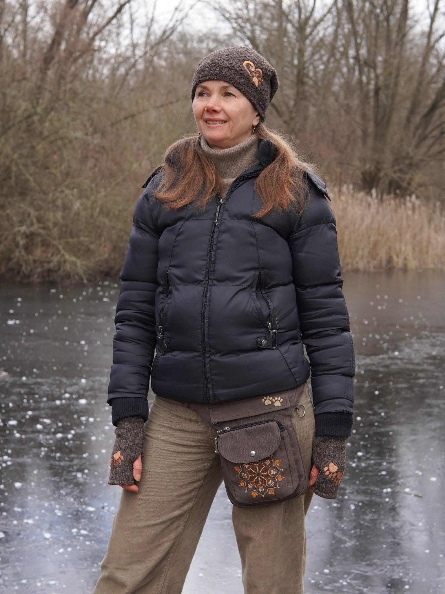 Ein Model präsentiert sitzend vor einem winterlich zugefrorenen See gestrickte Pulswärmer aus Schurwolle in Braun bestickt mit zwei Pfoten. Auf den Kopf trägt sie eine  Mütze aus Schurwolle Braun mit Herz & Pfotenstickerei.