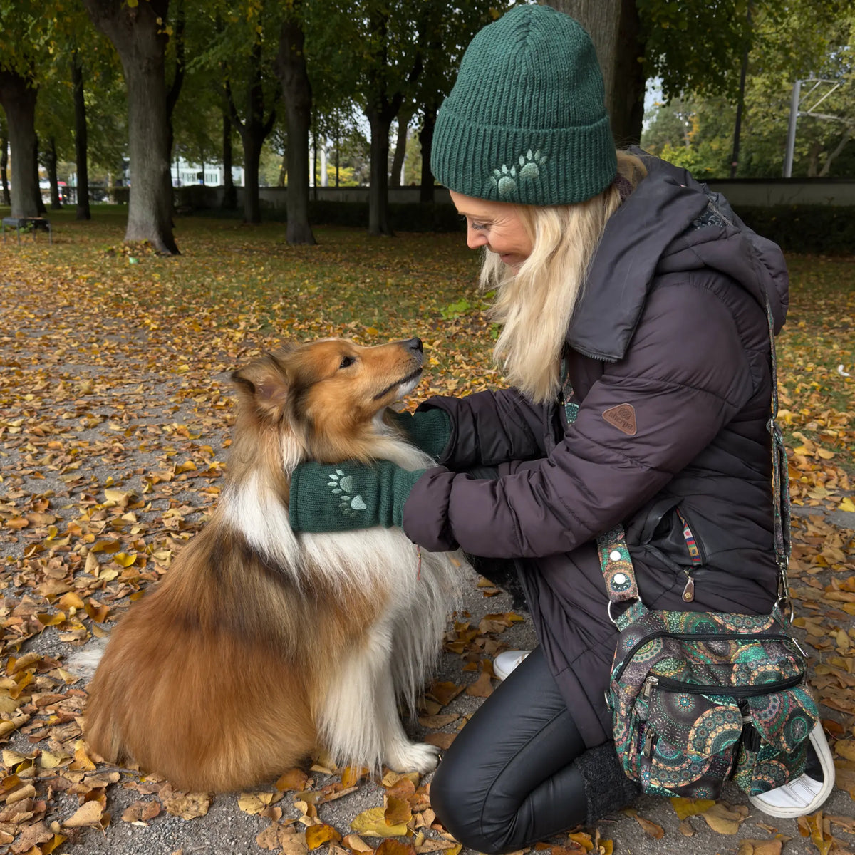 Frau mit Hund beim Herbstspaziergang, trägt grüne Nijens Beanie Tulsipur aus Schurwolle mit Pfotenstickerei, passende Armstulpen und bunte Gassitasche mit Mandala-Muster.