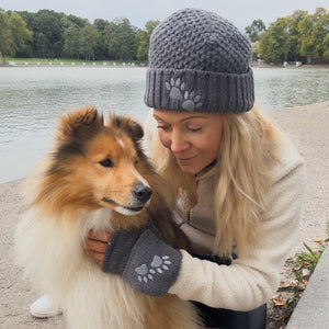 Frau trägt graue Wollmütze und Pulswärmer mit Pfotenmotiv von Nijens und kuschelt mit ihrem Hund am See – warm, gemütlich und stilvoll im Herbst.
