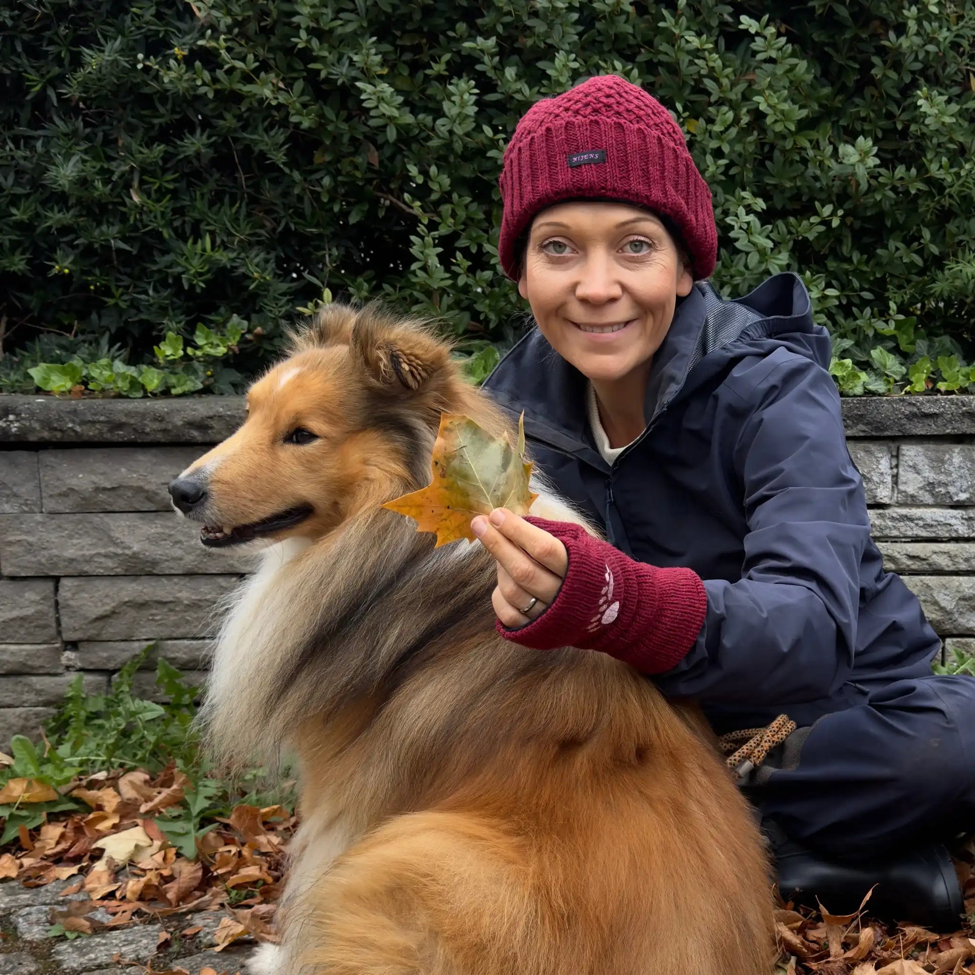 Frau mit bordeauxroter Nijens-Wollmütze und Pulswärmern mit Pfotenmotiv sitzt im Herbstlaub neben ihrem Hund – warme Accessoires für Spaziergänge im Herbst und Winter