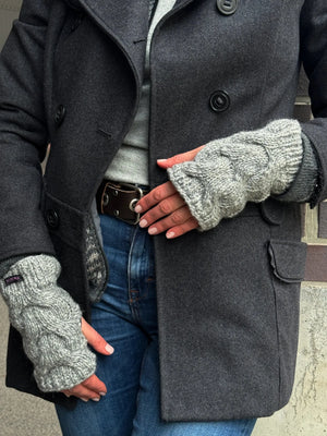 Nahaufnahme Hände: Model präsentiert handgestrickte Handstulpen, Pulswärmer aus Schurwolle grau, mit Daumenspalte und großem Zopfstrickmuster.