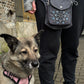 Stehendes Model präsentiert die Gassi-Tasche, Bauchtasche und Gürteltasche Barcelona 304 aus wasserabweisendem Nylonstoff in Grau mit Pfoten-Mandala-Stickerei. Vor ihr sitzt ein Hund. An den Händen trägt sie Handstulpen aus Schurwolle grau mit zwei Pfotenstickerei.