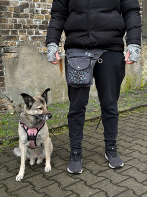 Ein Model in schwarz gekleidet, präsentiert die Gassi-Tasche, Bauchtasche und Gürteltasche Barcelona 304 aus wasserabweisendem Nylonstoff in Grau mit Pfoten-Mandala-Stickerei. Vor ihr sitzt ein Hund. An den Händen trägt sie Handstulpen aus Schurwolle grau mit zwei Pfotenstickerei.